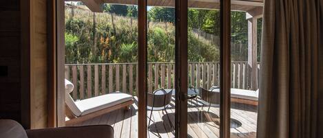 Una terraza de madera con un ventanal de vidrio que da a un jardín. Hay sillones de descanso y una mesa en la terraza.