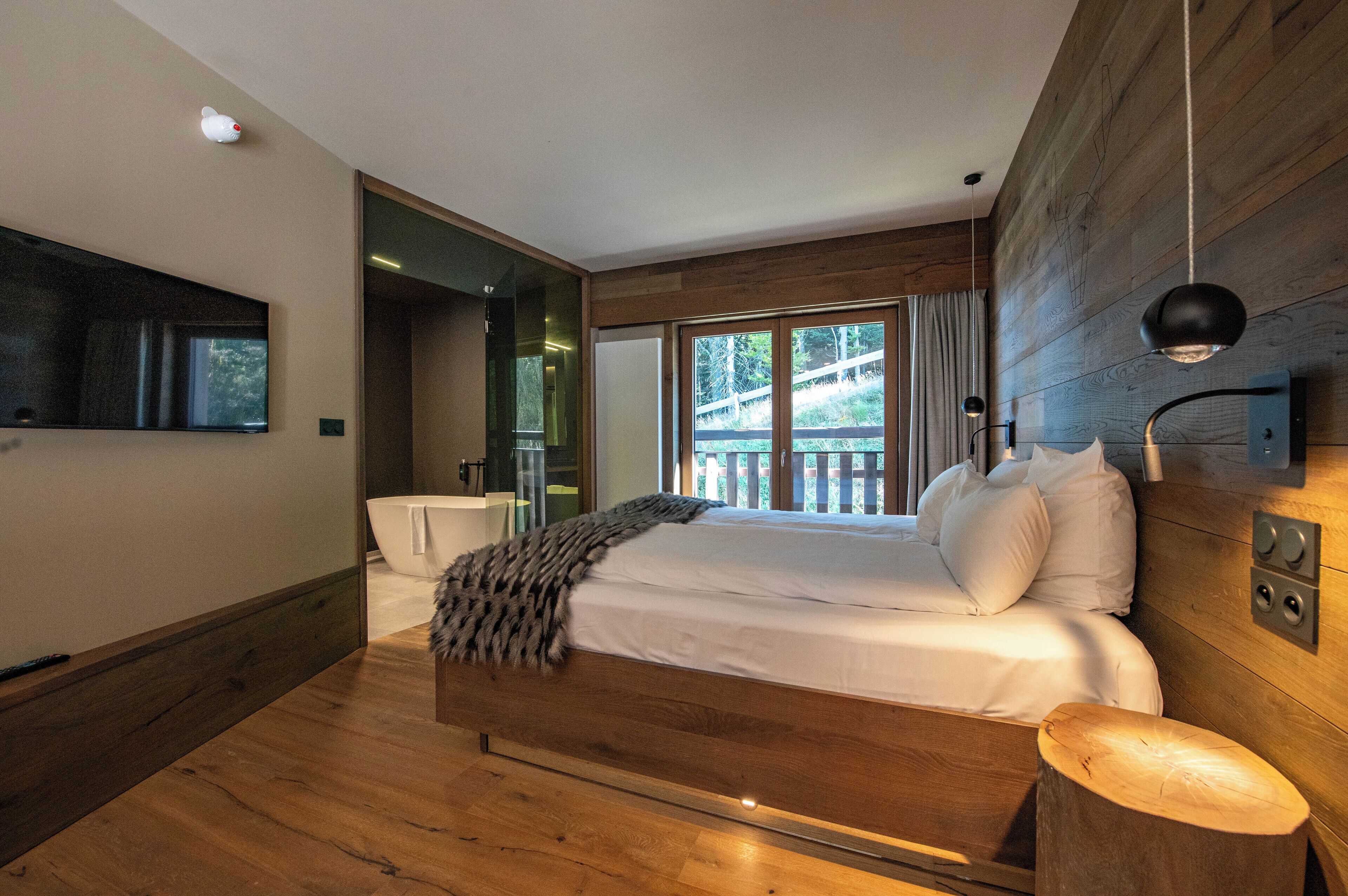 A modern bedroom with a large bed, wooden flooring, a wooden headboard, a large window with a view, and a mounted TV.