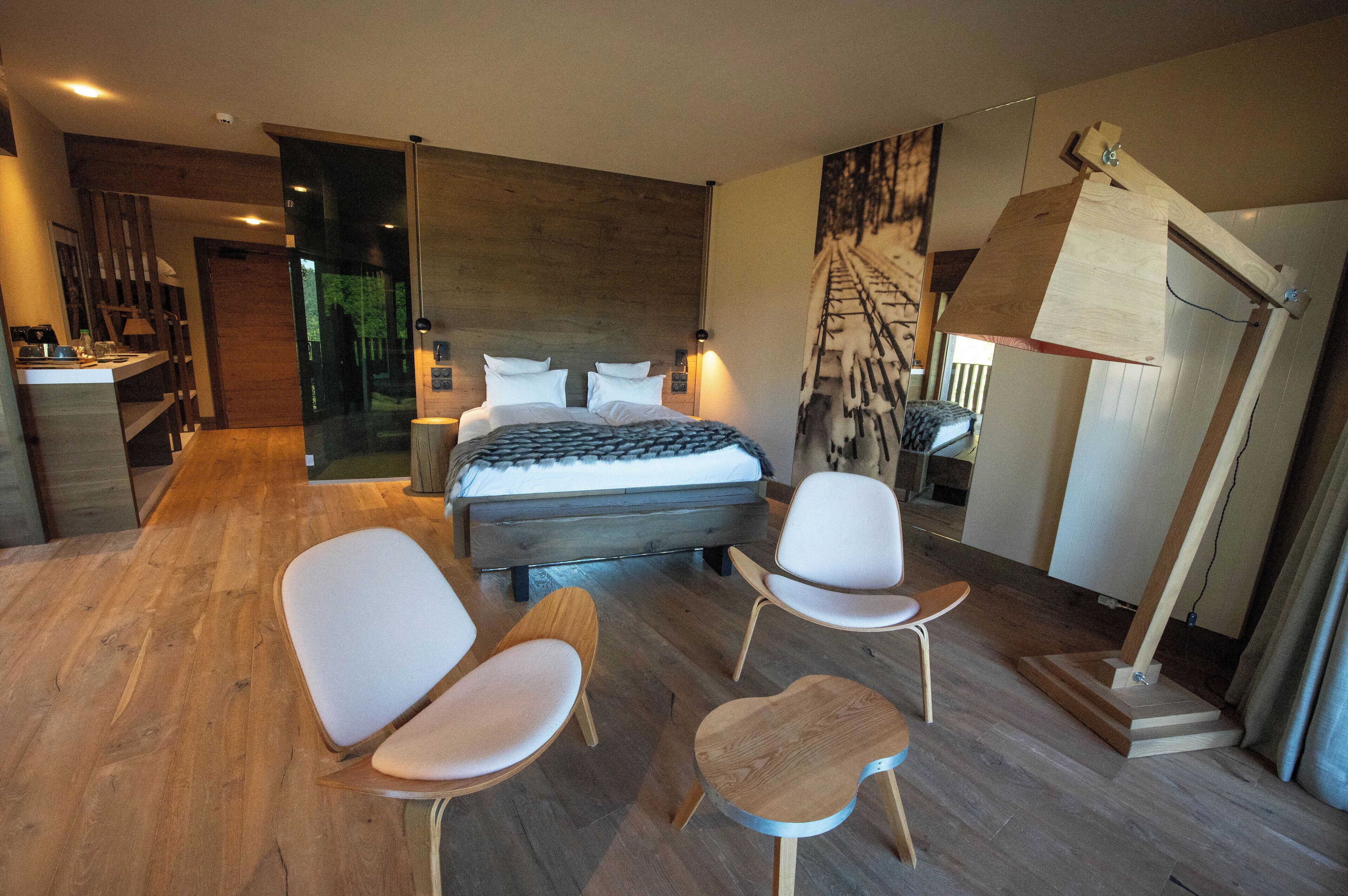 A modern bedroom with a large bed, two white chairs, a small wooden table, and a unique wooden ladder structure.