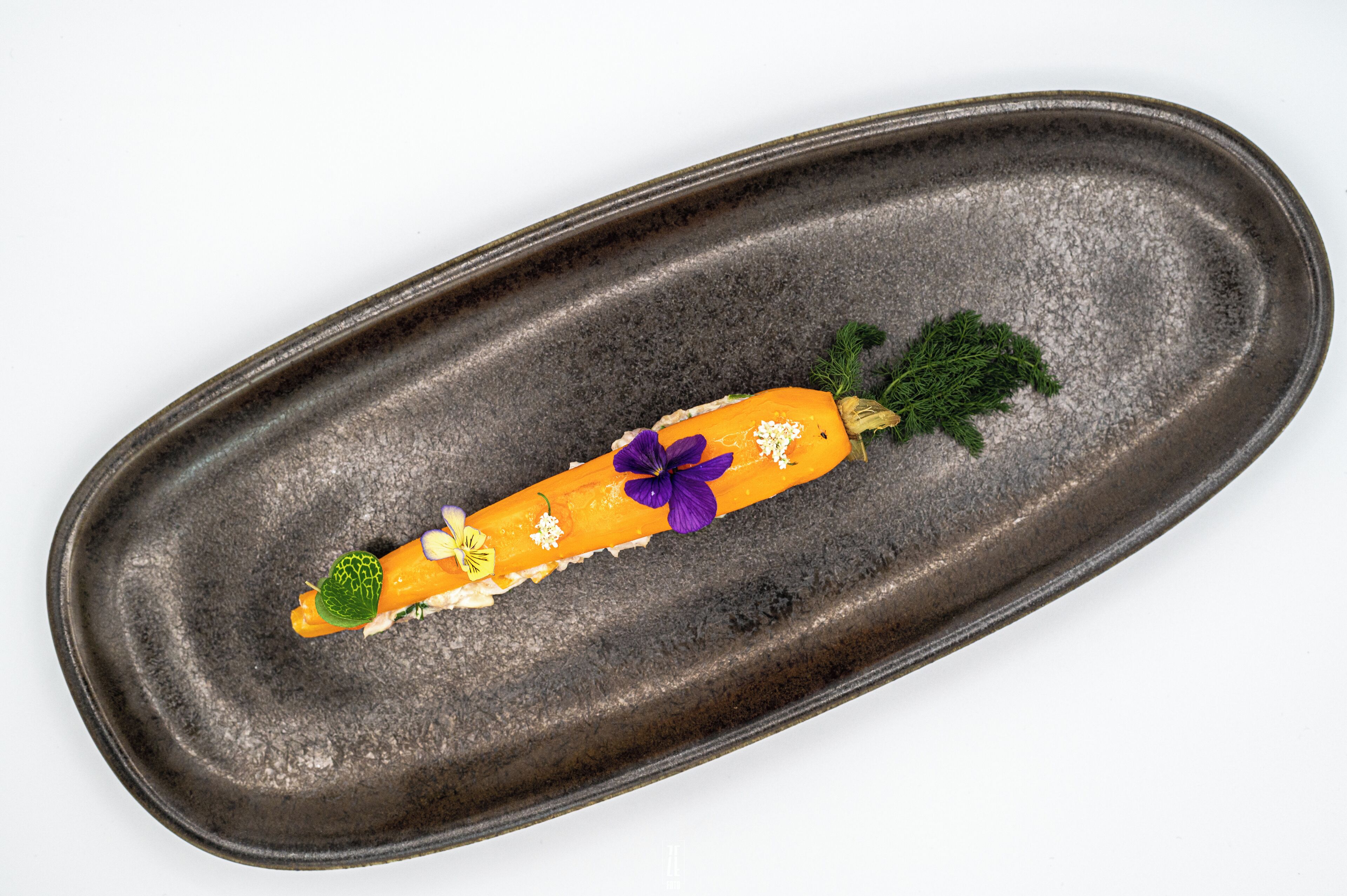 Un piatto scuro con una carota, fiori ed erbe aromatiche.