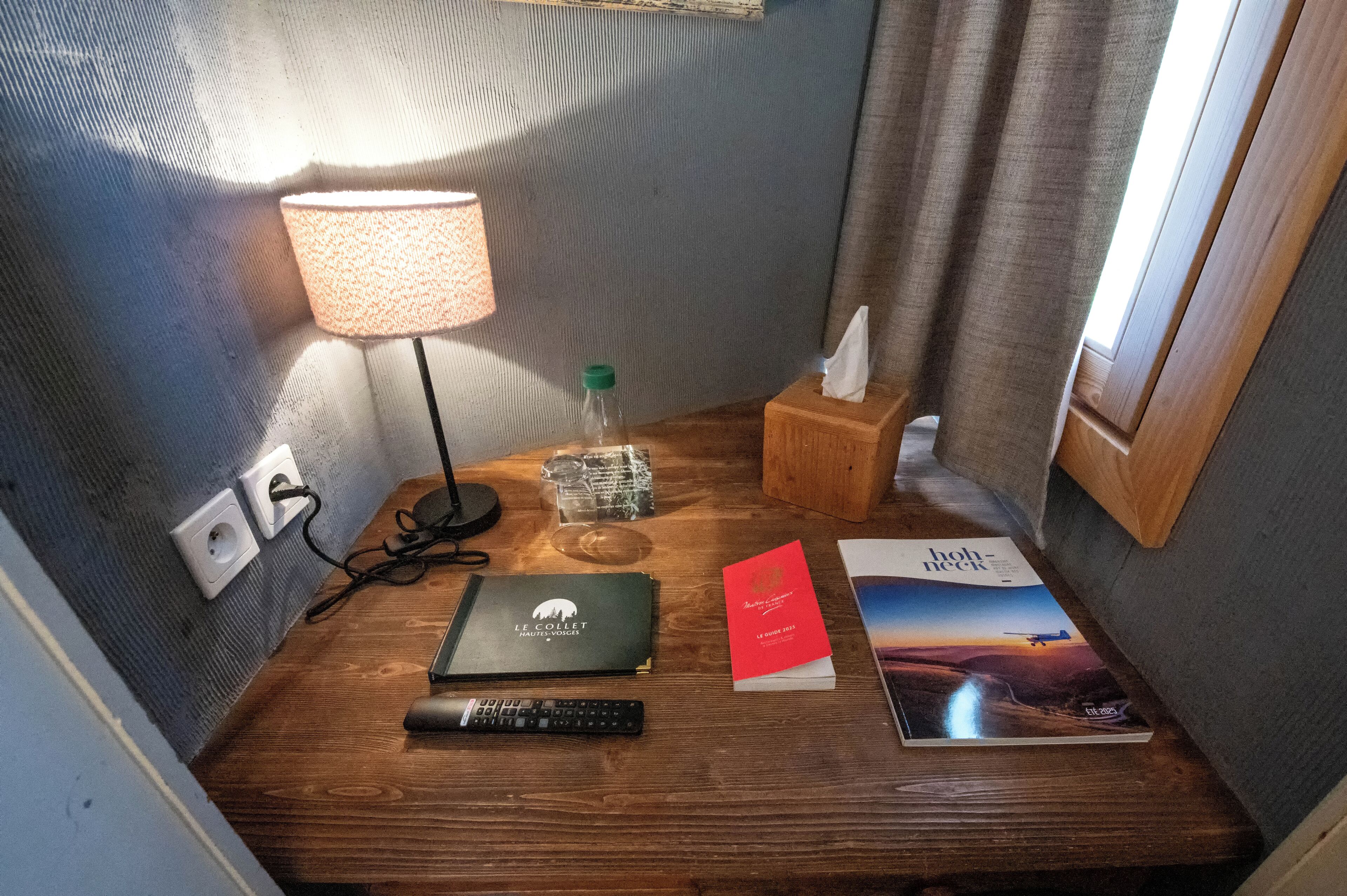 A neatly arranged hotel room with a wooden bedside table, a lamp, a remote control, a book, and a magazine.