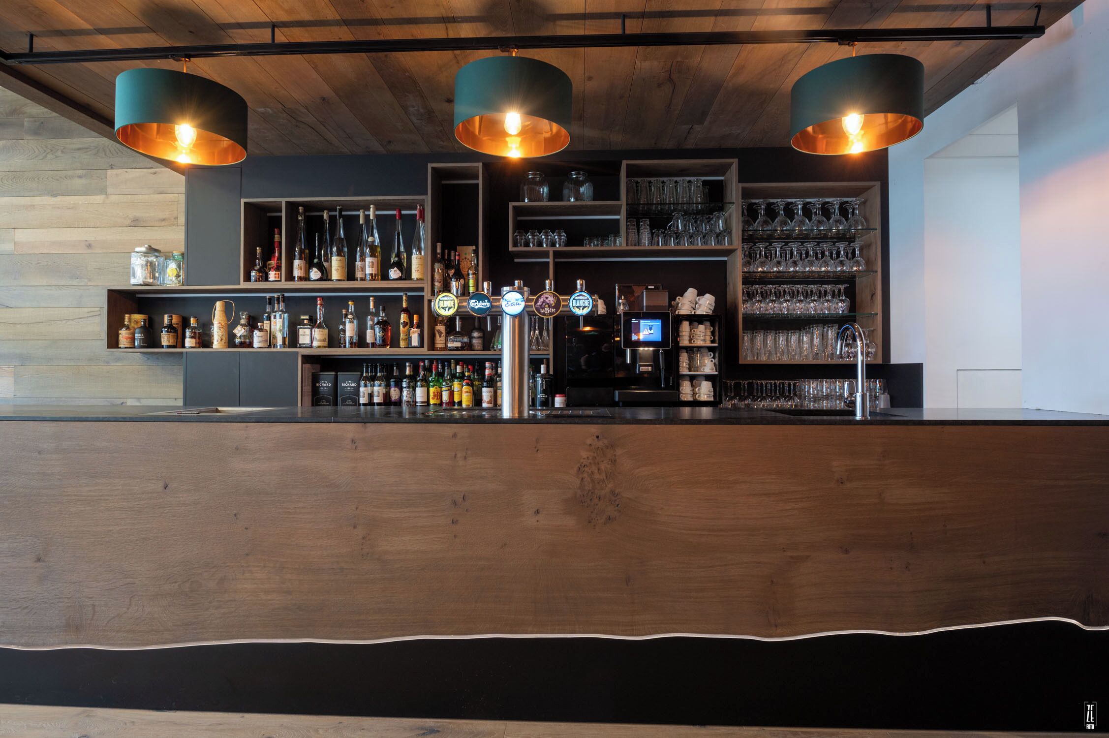 Una zona de bar con mostrador de madera, estantes con botellas y vasos, y tres lámparas colgantes.
