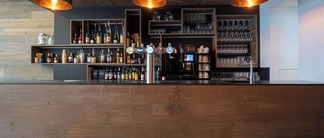 Una zona de bar con mostrador de madera, estantes con botellas y vasos, y tres lámparas colgantes.