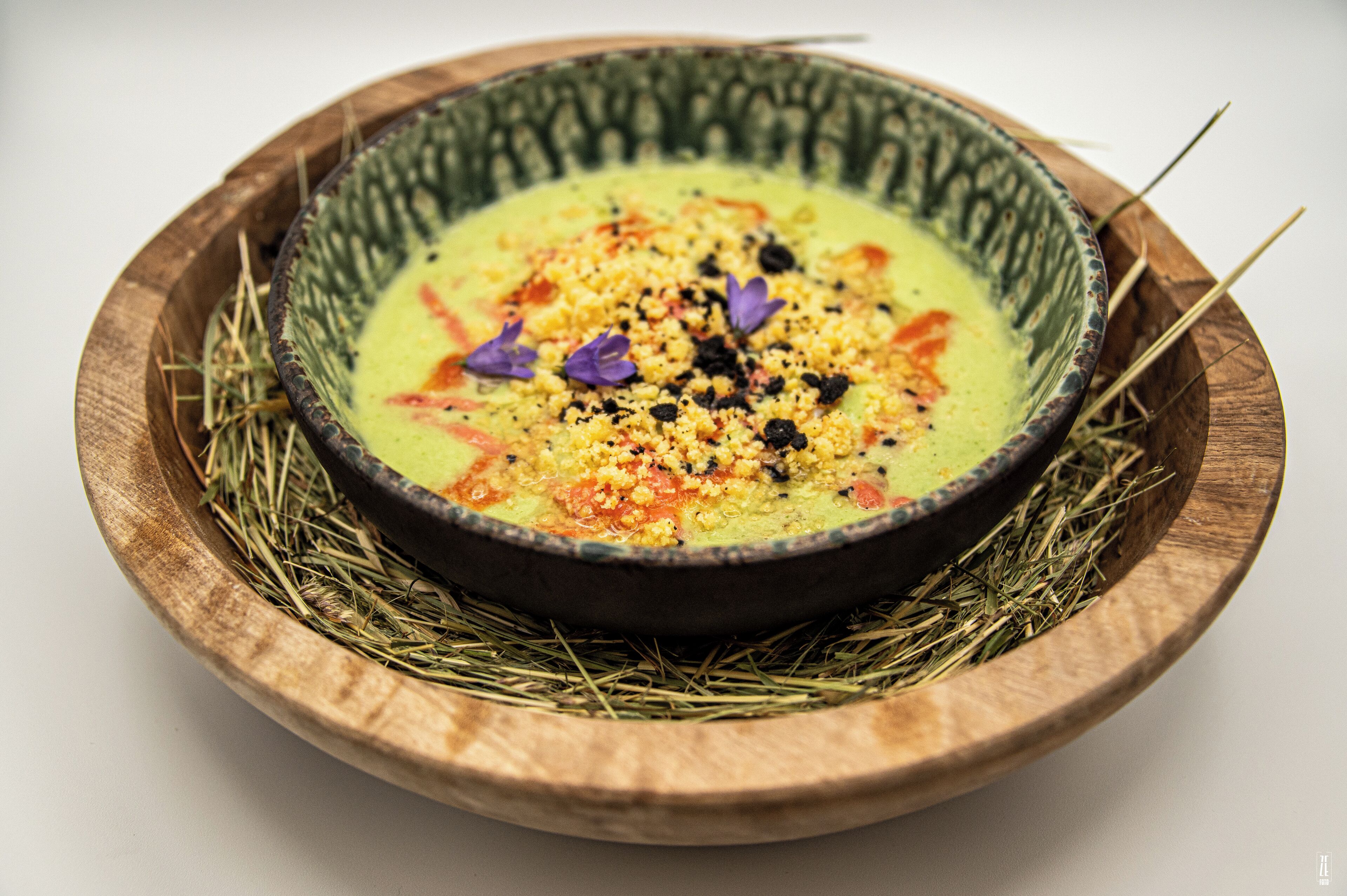 En skål med soppa, garnerad med örter och ätbara blommor, serverad i en rustik träskål på en bädd av halm.