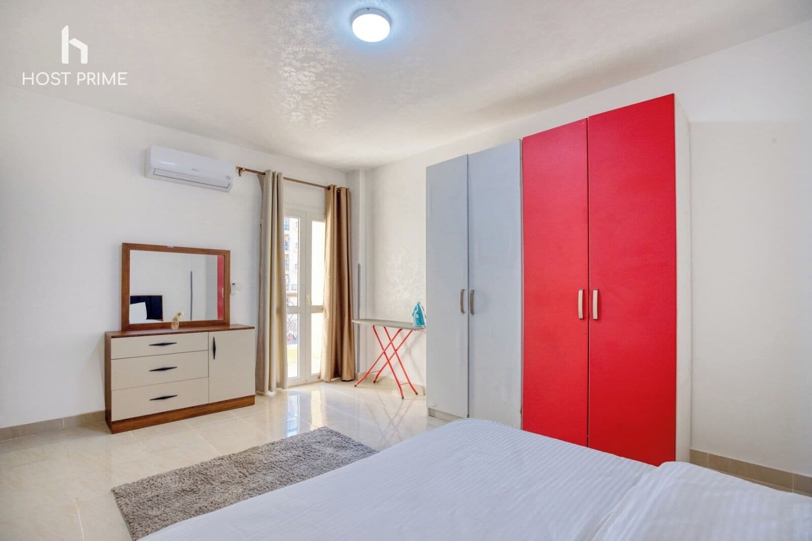 Quarto com cama, cômoda, guarda-roupa vermelho, espelho e janela com cortinas.