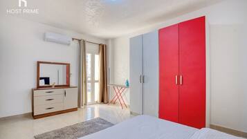 Quarto com cama, cômoda, guarda-roupa vermelho, espelho e janela com cortinas.