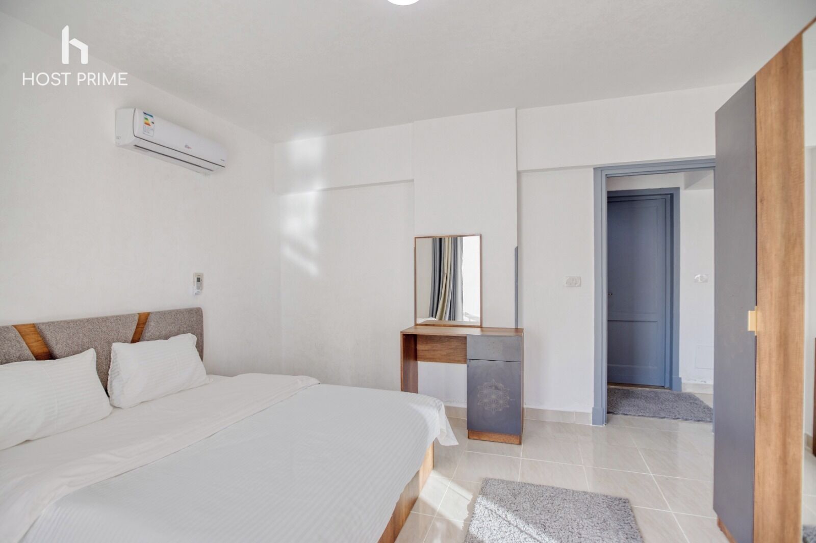 A modern hotel room with a large bed, a wooden nightstand, a mirror, and a door leading to another room.