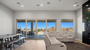 A modern living room with a sectional sofa, a dining table, and large windows offering a view of the desert landscape. - Four Paws & Fairways | Golf Course Front | Pet Friendly | Over $500 in complimentary activities (Mesquite)