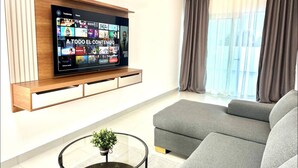 A modern living room with a flat-screen TV mounted on a wooden shelf, grey sofas, a glass coffee table with a plant, and a large window with curtains. - Modern Rooftop Penthouse Private Jacuzzi & View (Santiago de los Caballeros)