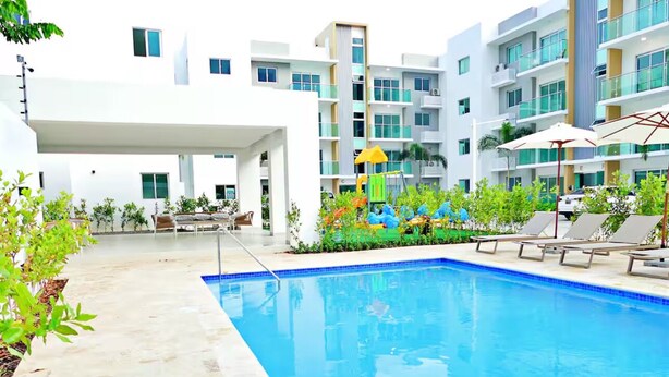 Una piscina con sillones de descanso y sombrillas, rodeada de vegetación y edificios modernos.