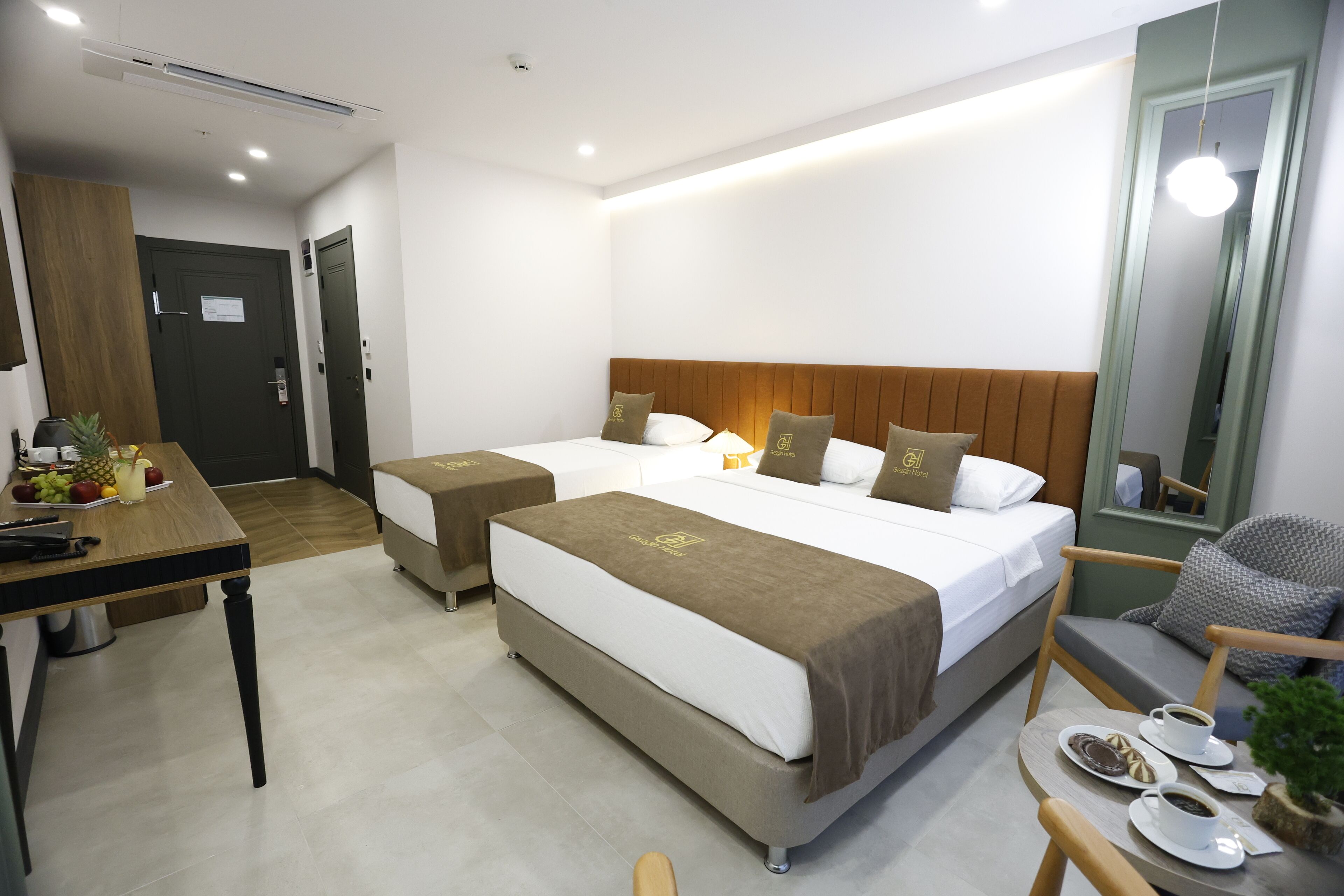 A hotel room with two beds, a desk with a fruit bowl, a coffee table with cups and a plant, and a mirror.