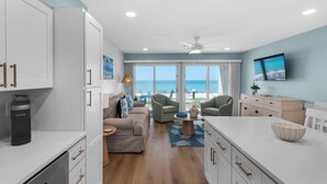 Una cocina moderna con armarios blancos y una isla grande, una sala de estar con sofá y sillones, y vistas a la playa a través de amplias ventanas.