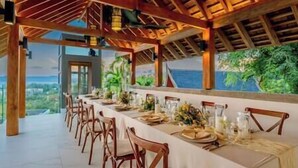 A long dining table set for a meal with chairs, under a wooden ceiling with hanging lights, and a view of the outdoors. - Zenith Villa | Luxury - 6 Bedroom Villa in Surin with Sea View (Cheong Thale)