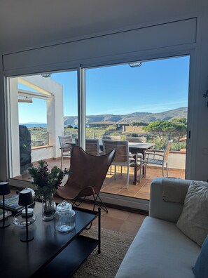 A modern living room with a large sliding glass door leading to an outdoor patio. The room features a sofa, a dining table, and a chair. - Waterfront apartment (Cadaqués)