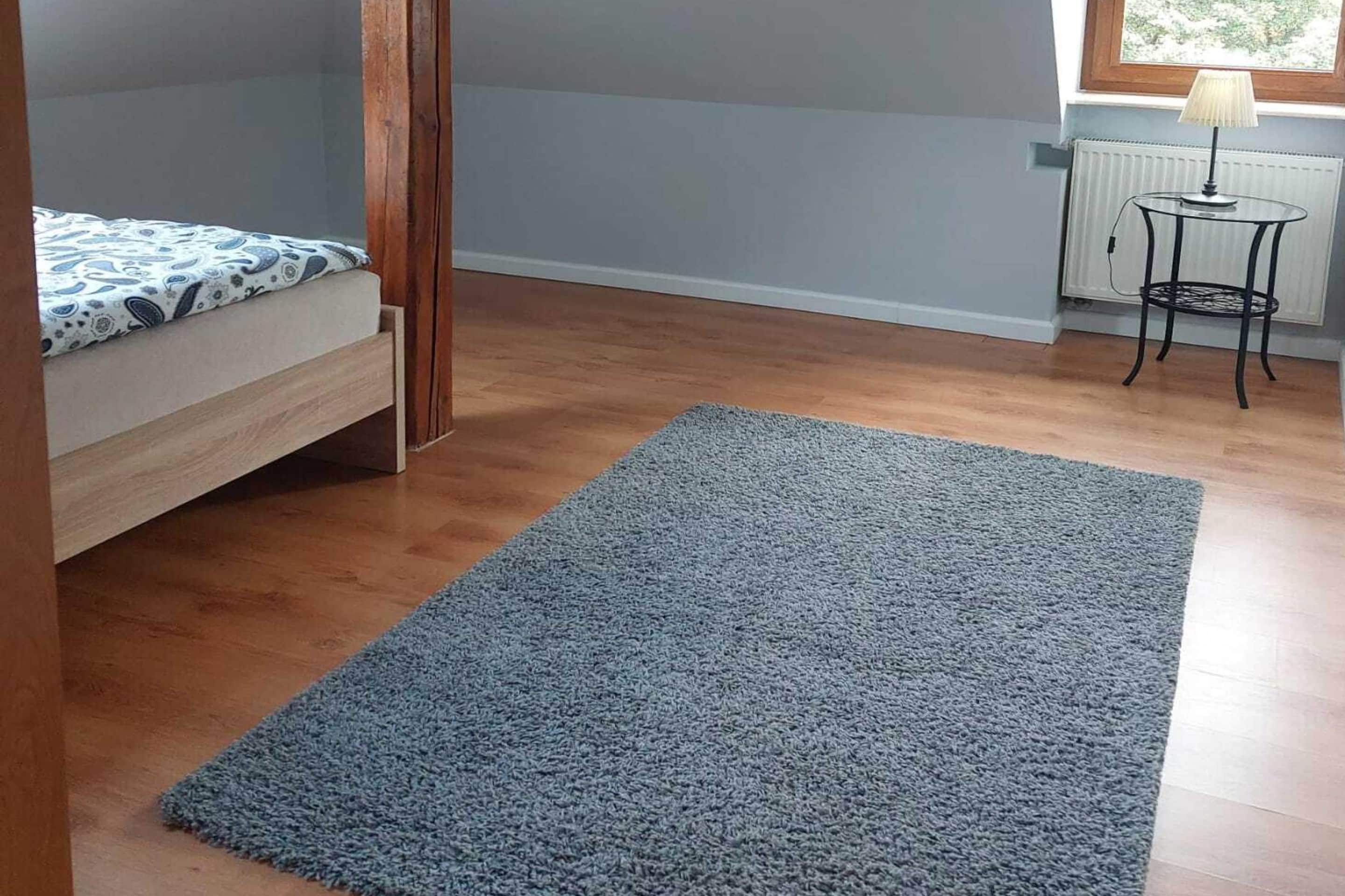 Een slaapkamer met een bed, een grijs vloerkleed, een kleine tafel met een lamp en een raam.