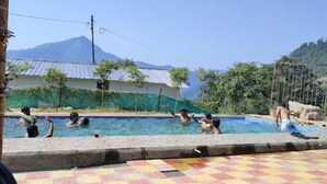 A swimming pool with people enjoying the water, surrounded by greenery and a mountain in the background. - Shivadri Resort  (Rishikesh)