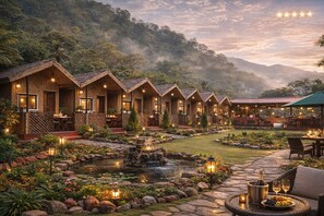 A row of wooden cabins with outdoor seating, a garden with a pond, and a mountain backdrop. - Shivadri Resort  (Rishikesh)