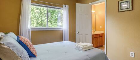Une chambre à coucher avec un lit, des oreillers, une fenêtre avec des rideaux, une porte donnant sur une salle de bain et un tableau encadré au mur.