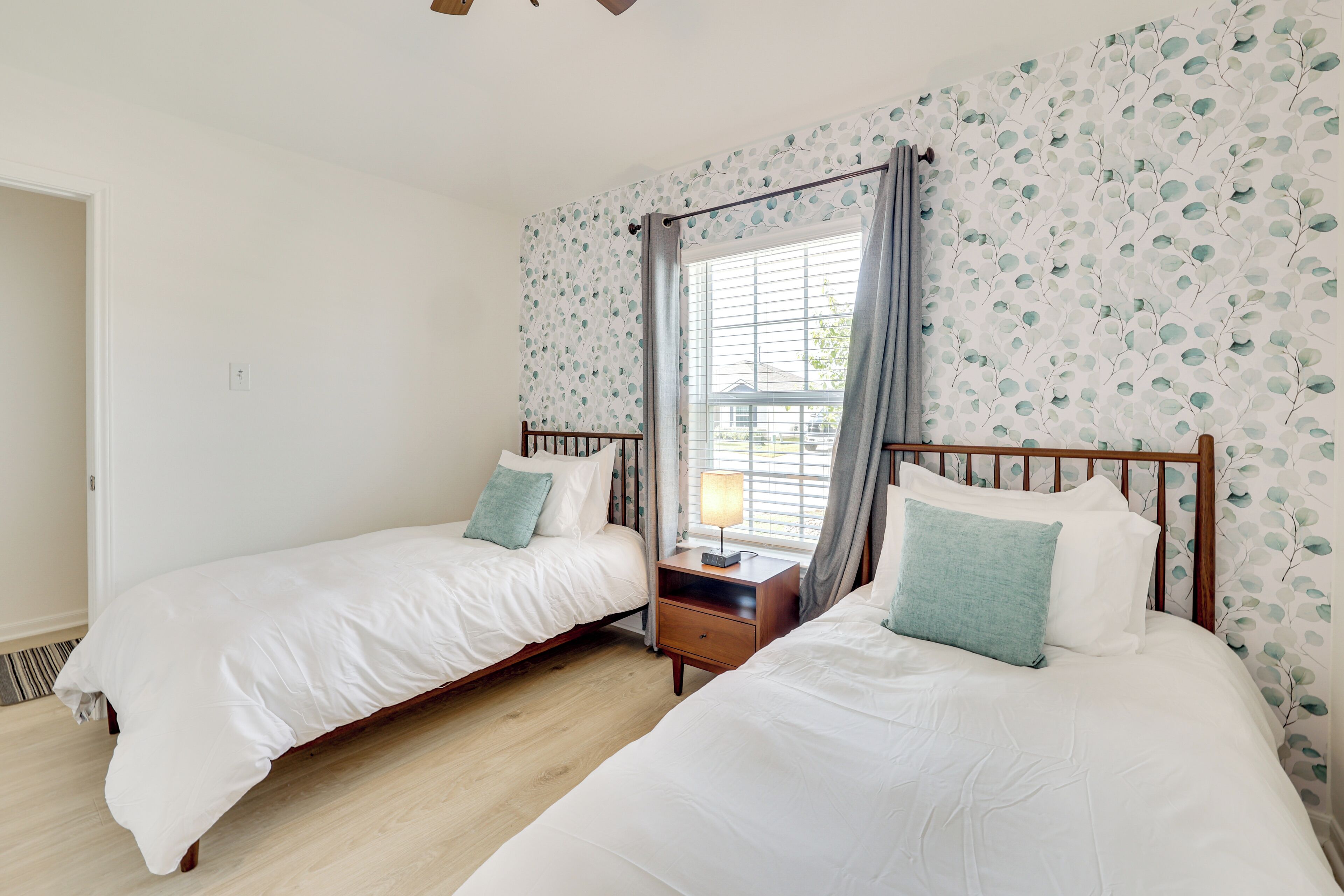 A bedroom with two single beds, a nightstand with a lamp, a window with curtains, and a patterned wall.
