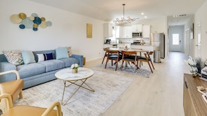 A modern living room with a blue sofa, a round coffee table, and a dining area with wooden chairs and a white table. - Game Room + Private Yard: Modern San Antonio Home (San Antonio)