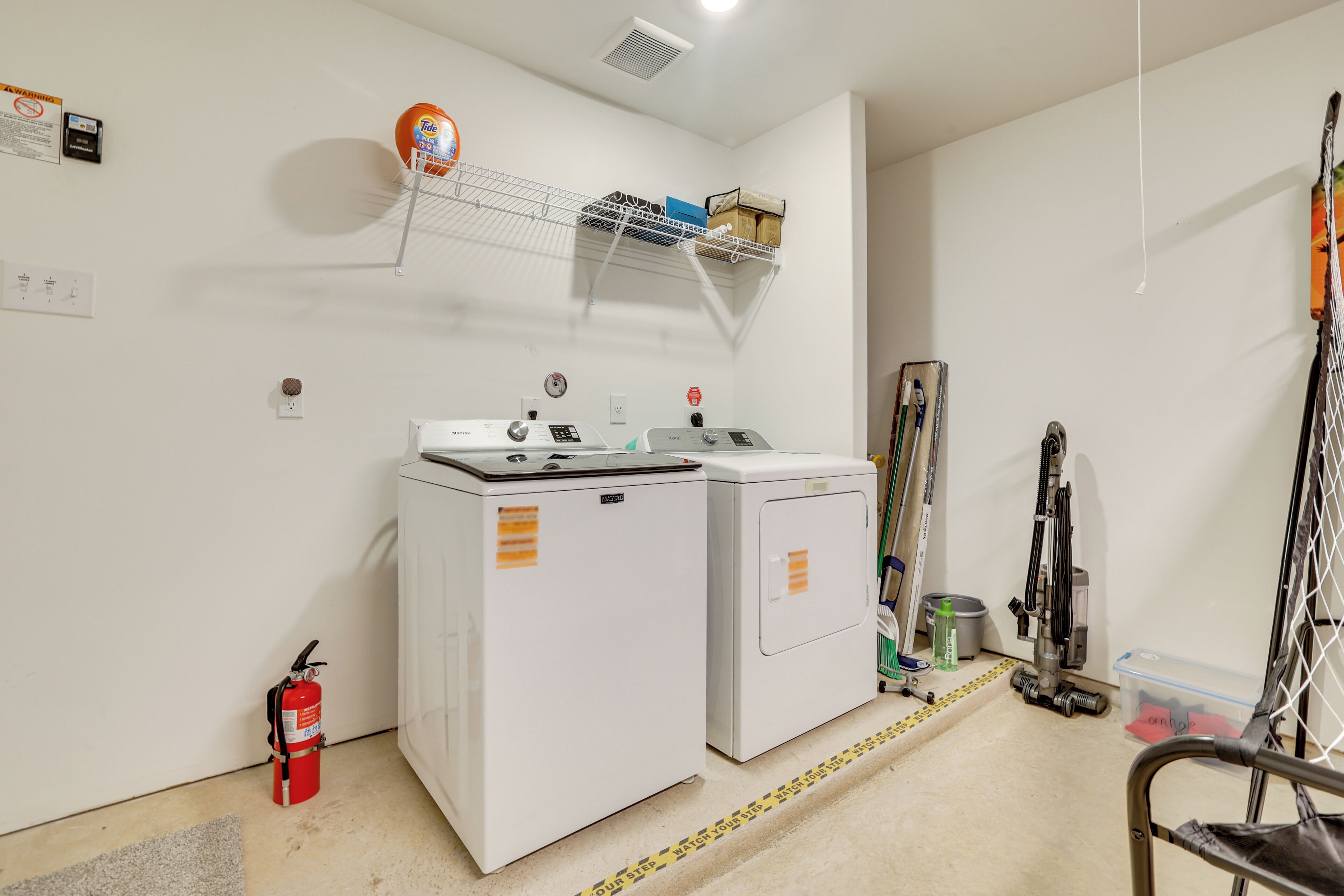 A laundry room with two washing machines, a fire extinguisher, a shelf with cleaning supplies, and a rack with skis.