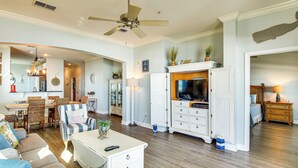 Amplia sala de estar con ventilador de techo, comedor con mobiliario de mimbre, cocina con televisor y dormitorio con cama y cómoda.