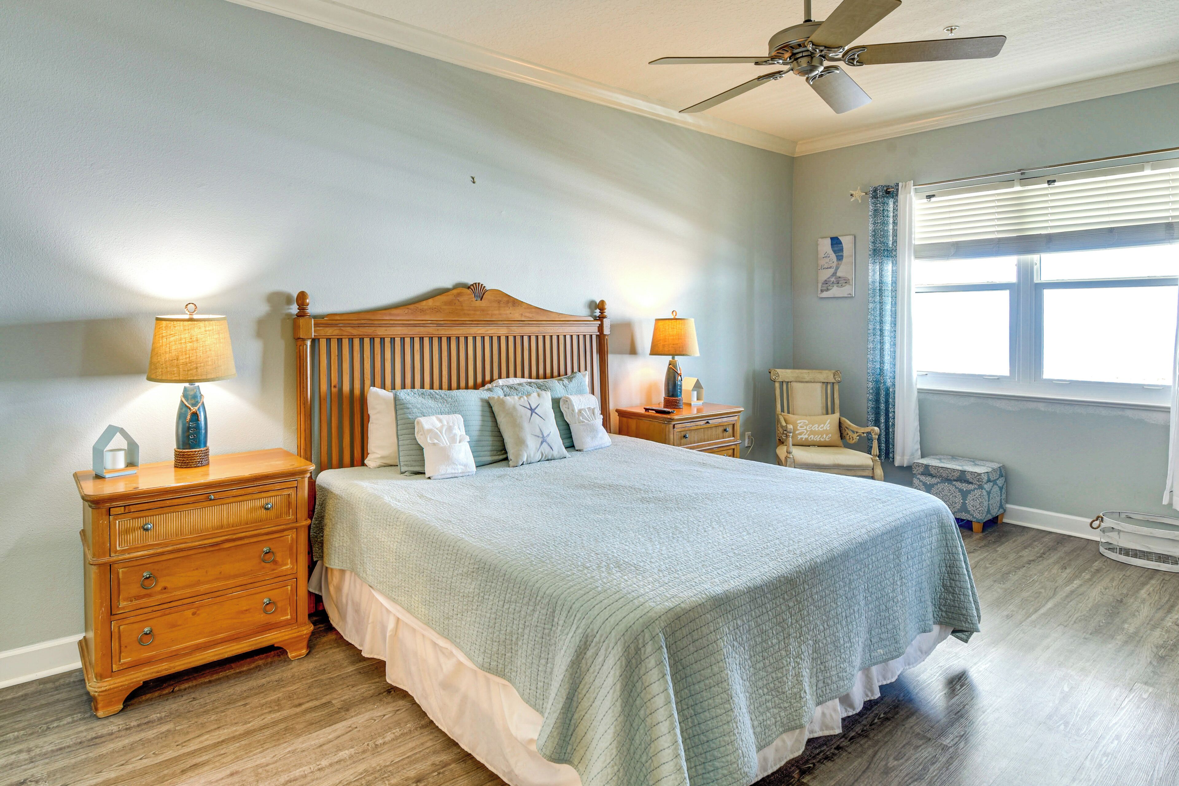 A bedroom with a large bed, two bedside tables, a ceiling fan, a window with blinds, and a small chair.