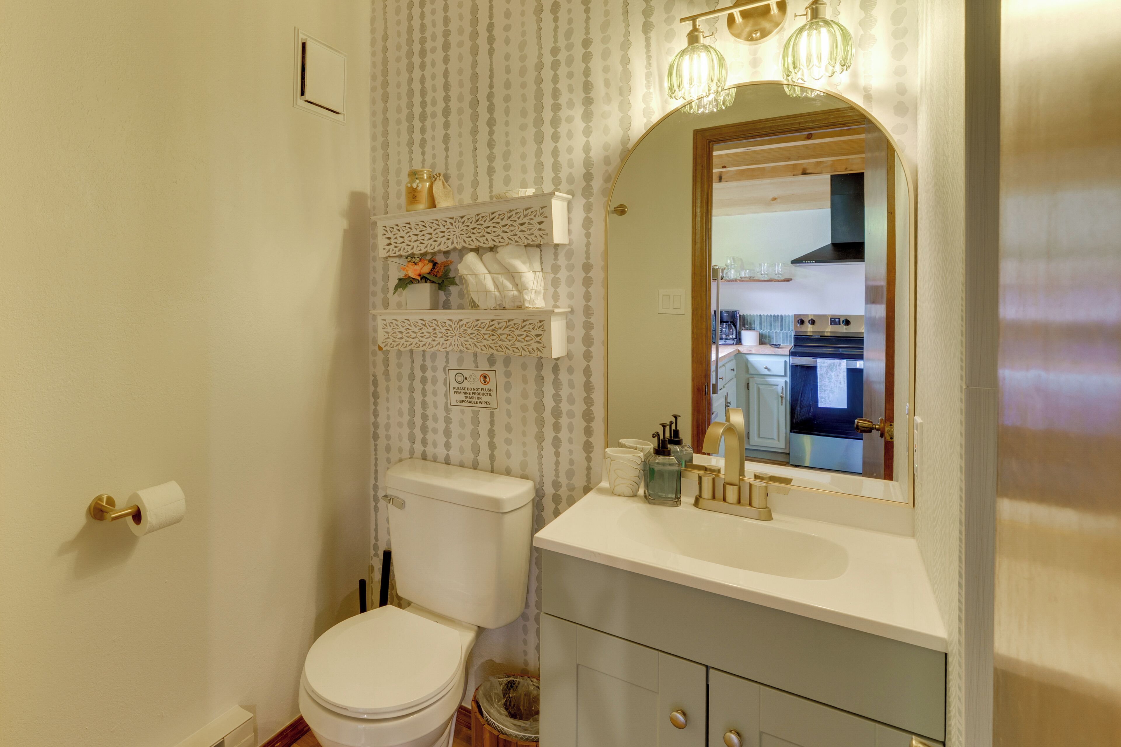 A bathroom with a toilet, sink, and mirror. There are shelves above the toilet and a trash can next to it.
