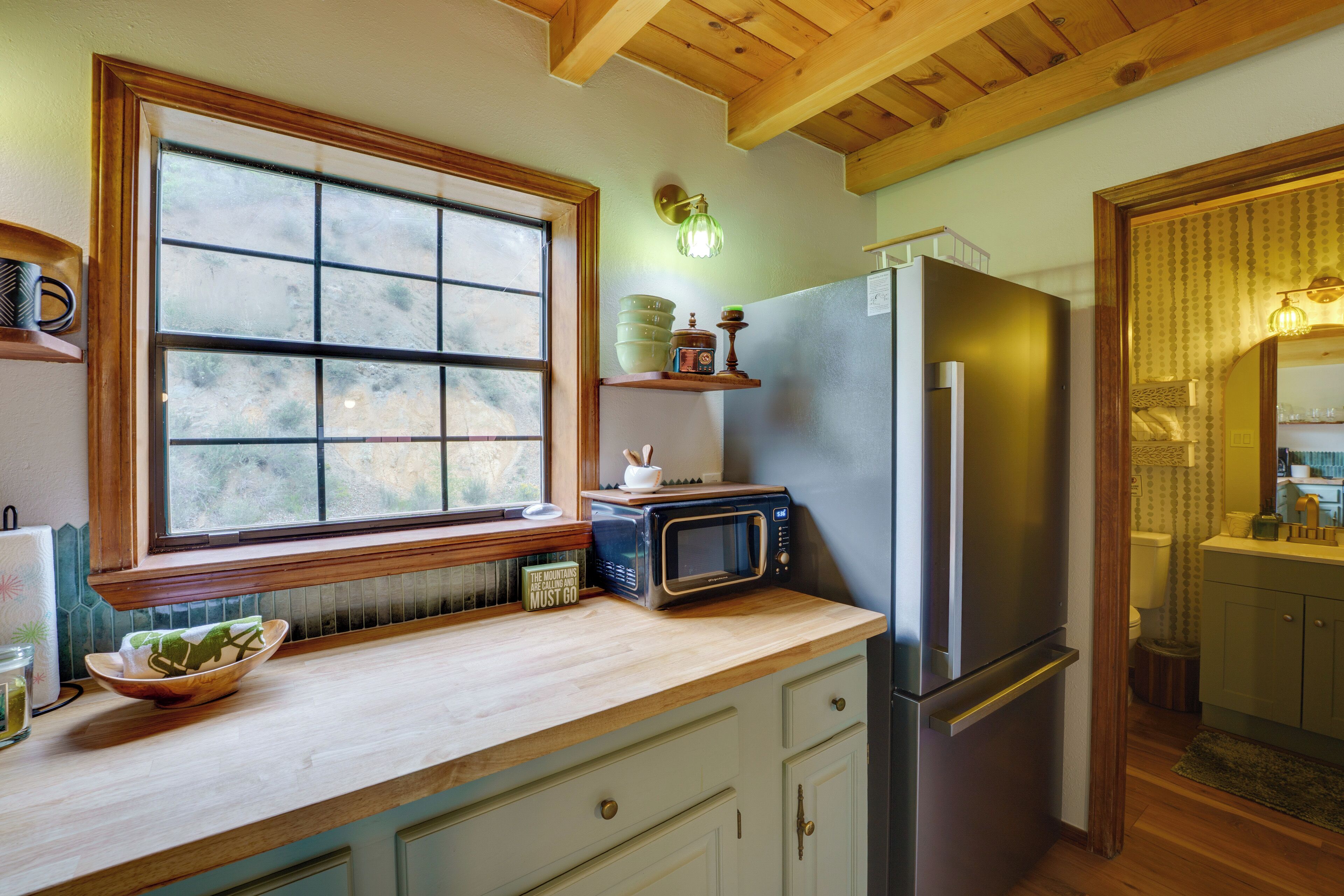A kitchen with a wooden countertop, a stainless steel refrigerator, a microwave, and a window with a view of greenery.