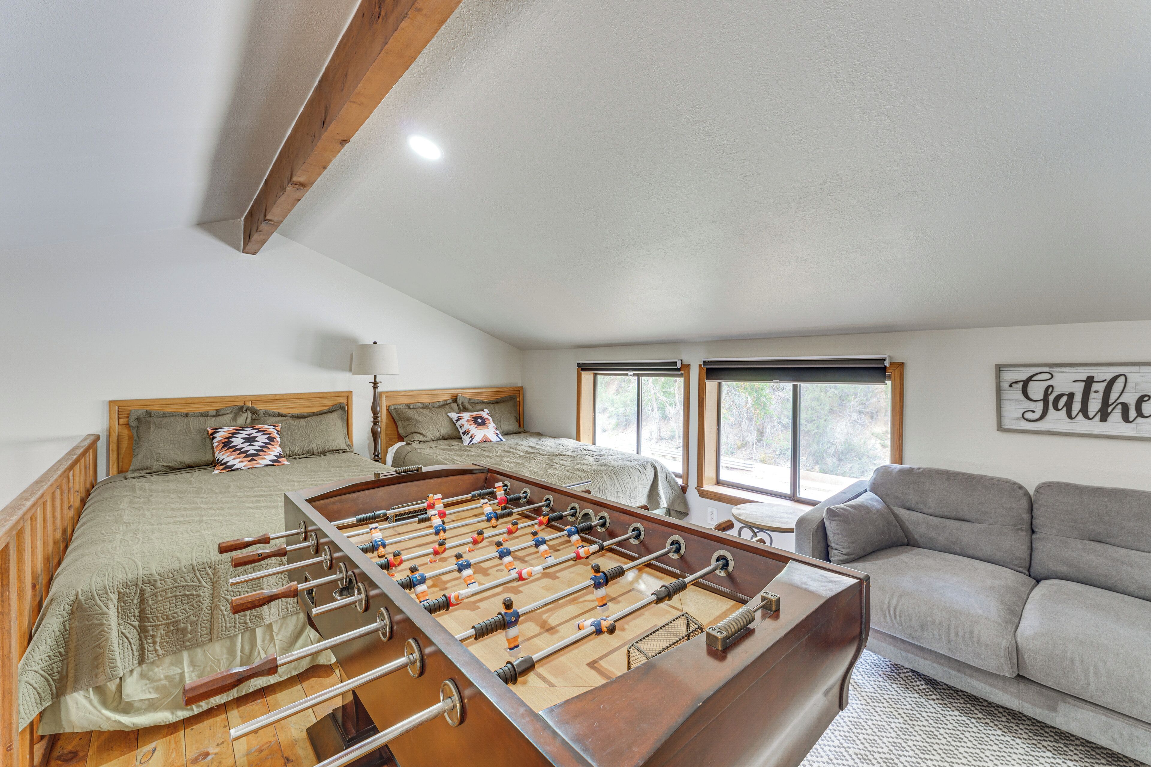 A cozy room with a foosball table, a bed with a wooden headboard, a gray sofa, and a window with a view of trees.