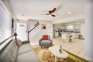 A modern living room with a grey sofa, a small table, and a dining area with a white table and chairs. A ceiling fan is visible, and there is a staircase leading to another level.
