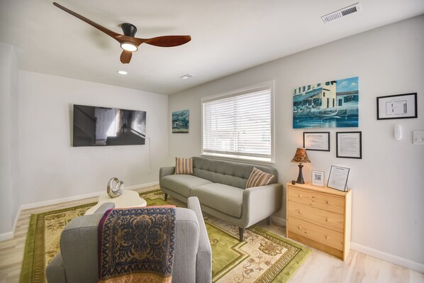 A living room with a grey sofa, a wooden side table, a ceiling fan, and a TV mounted on the wall.