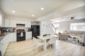 A modern kitchen with white cabinets, a black stove, and a refrigerator. There is a dining table with chairs and a sofa in the living area.