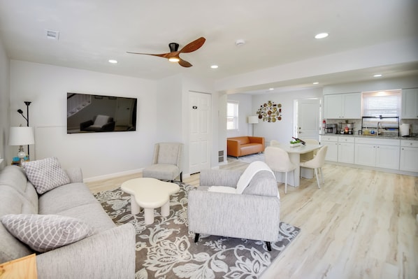 A spacious living room with a grey sofa, a white armchair, and a coffee table. There is a ceiling fan, a TV mounted on the wall, and a kitchen area visible through an open door.