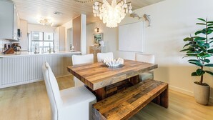 A dining area with a wooden table, white chairs, and a bench. There is a potted plant on the right.