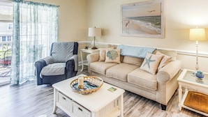 A living room with a beige sofa, a blue armchair, a coffee table with a decorative tray, and a painting of a beach scene on the wall.