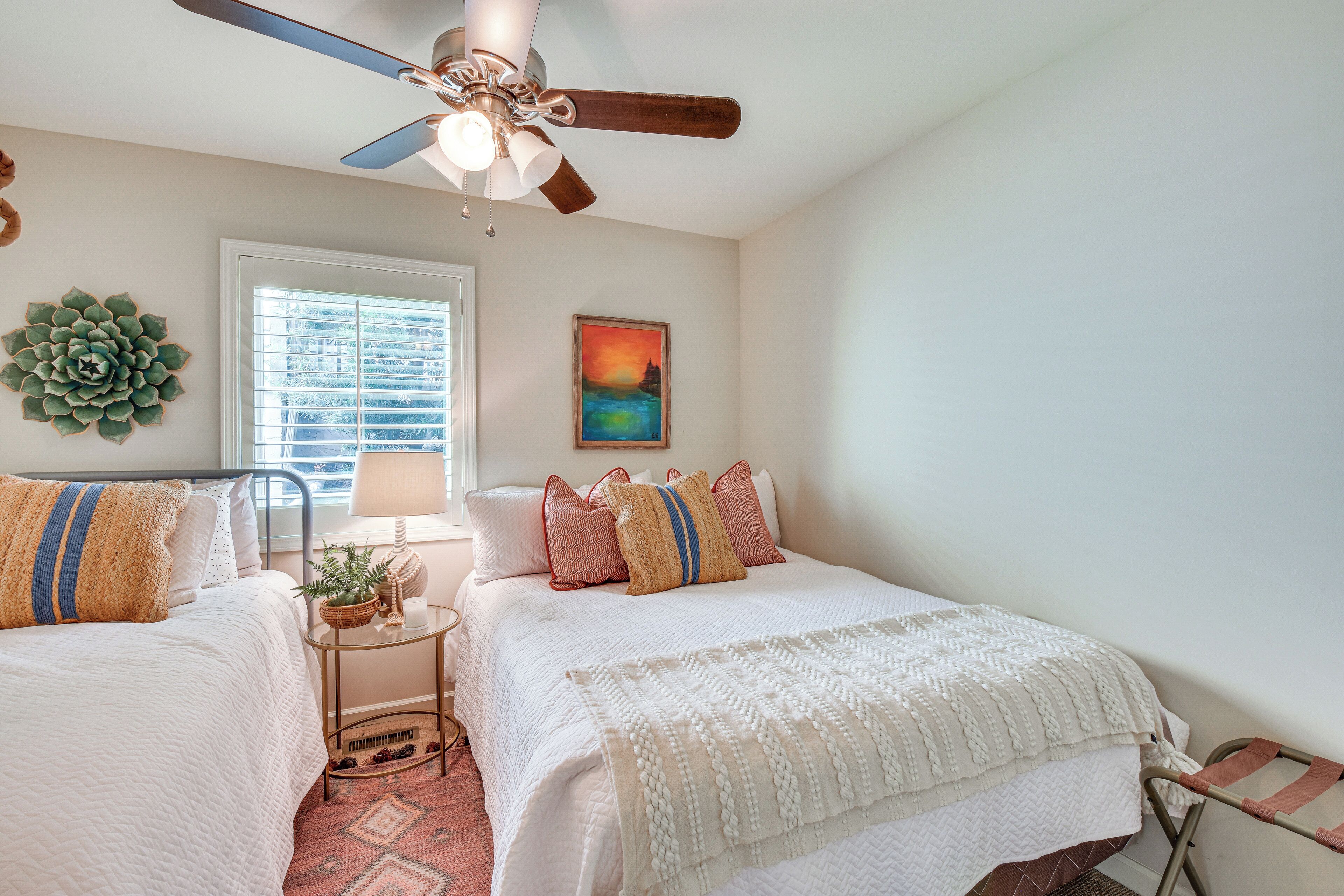 A bedroom with two beds, a ceiling fan, a window with blinds, a painting, and a decorative plant.