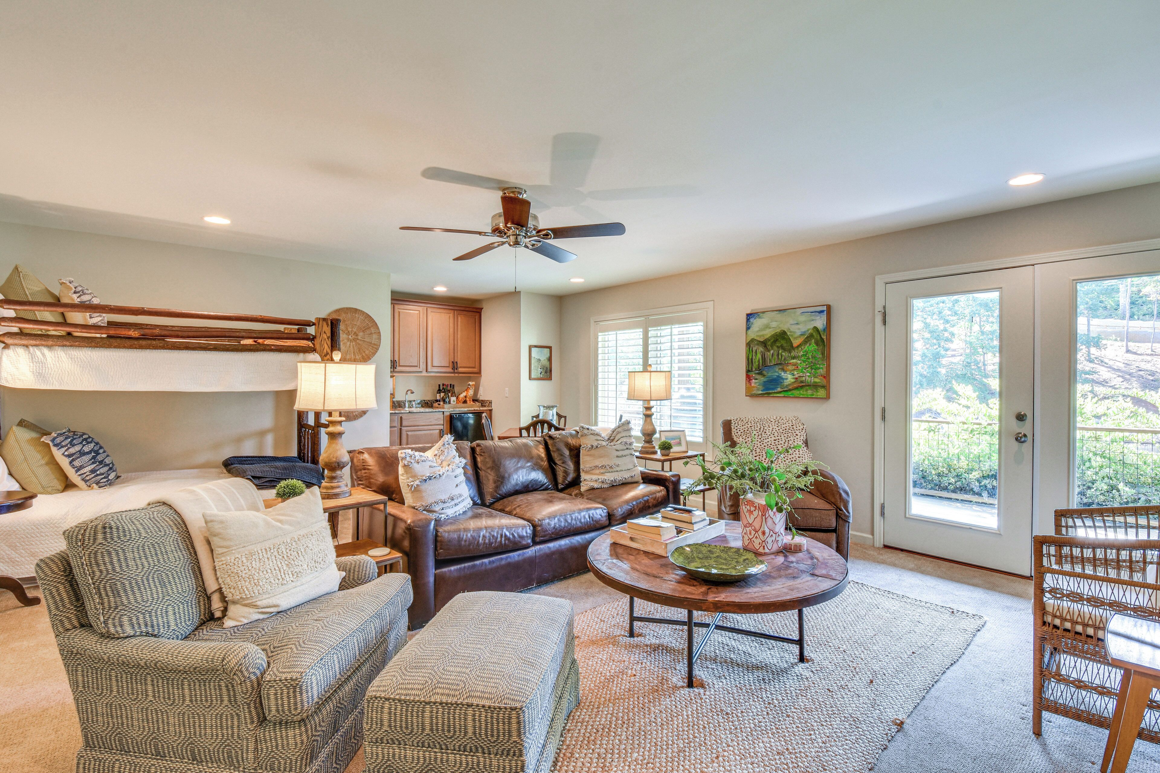 A spacious living room with a brown leather sofa, a green patterned sofa, and a wicker chair. There's a wooden coffee table with a bowl and a lamp. A ceiling fan is visible, and the room has large windows with a view of greenery outside.