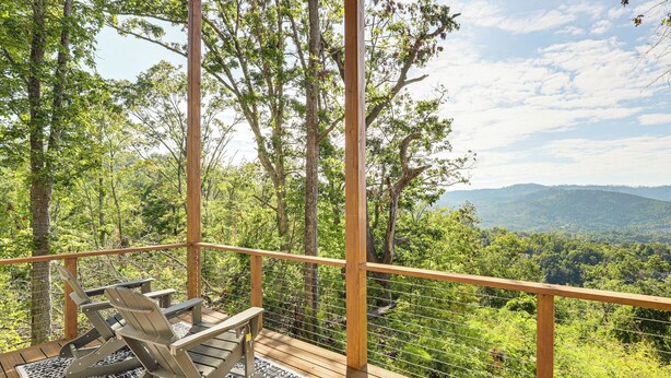Una terraza de madera con dos sillas Adirondack y un tapete estampado, con vistas a un paisaje boscoso y montañas a lo lejos.
