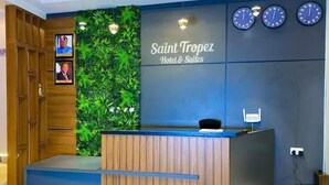 A reception desk with a wooden finish, a green wall with foliage, and clocks on the wall. - Saint Tropez Hotel and Suites (Abuja)