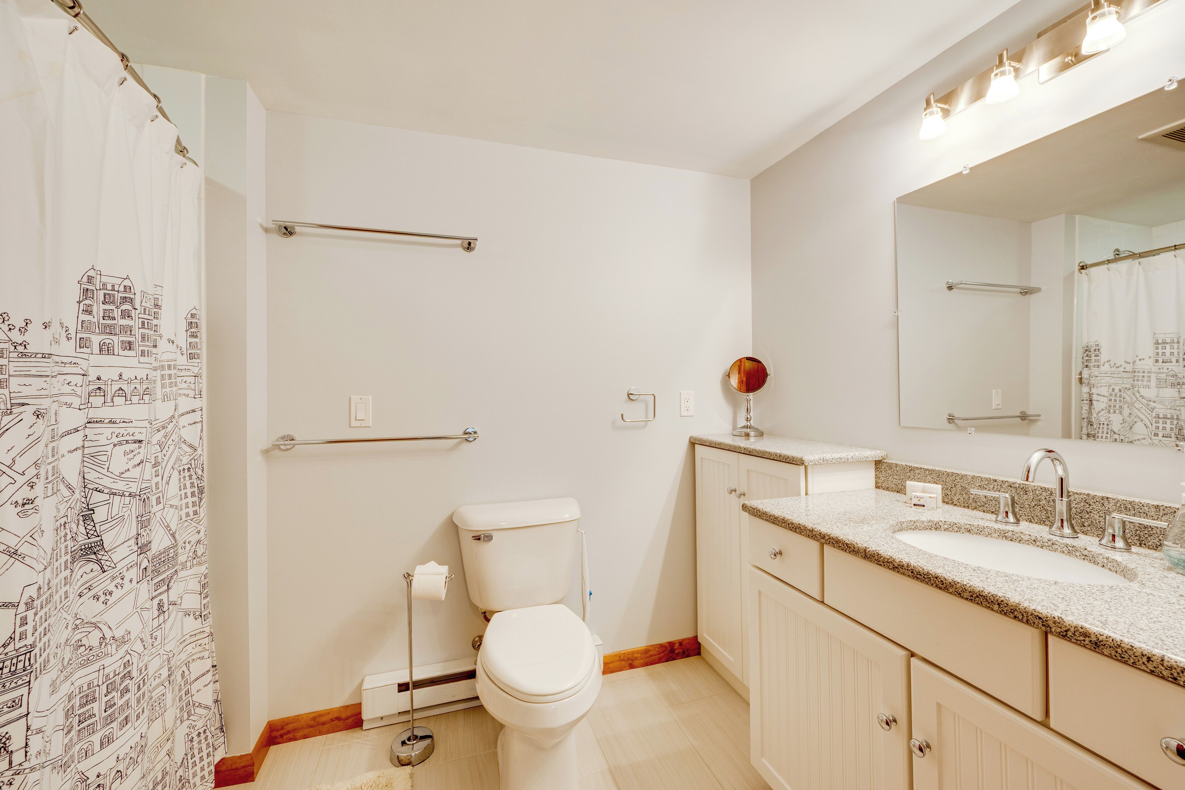 Une salle de bain équipée de toilettes, d’un lavabo, d’un miroir et d’un rideau de douche orné d’un motif représentant un paysage urbain.