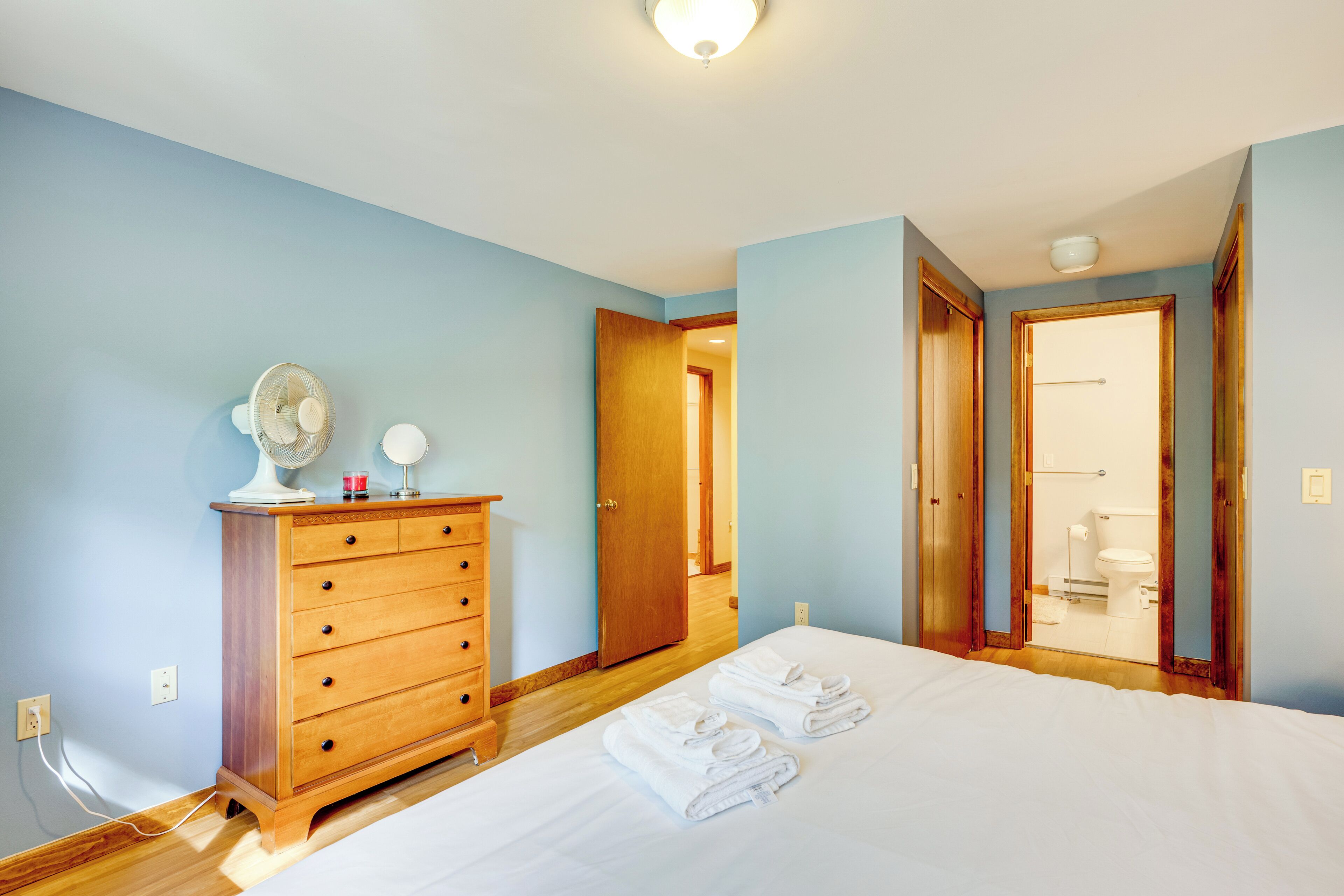 Un dormitorio con un aparador de madera, una cama con ropa blanca y un baño visible a través de una puerta abierta.