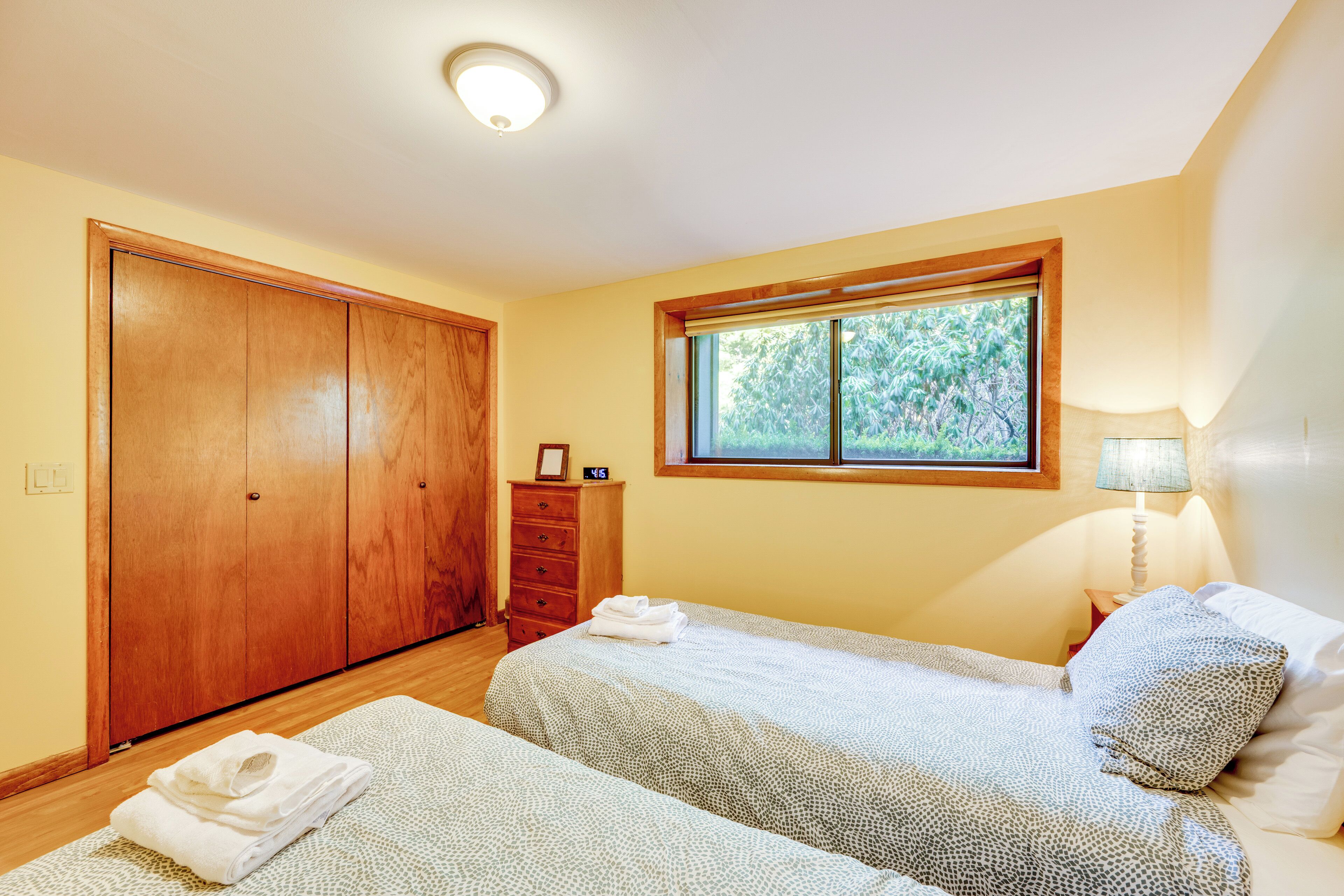 A bedroom with two beds, a wooden wardrobe, a window with a view of trees, and a bedside lamp.