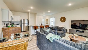 A modern living room with a grey sofa, a coffee table, and a TV. There is a kitchen area with white cabinets and a refrigerator. - Screened Patio: Townhome 3 Mi to Dtwn Wilson! (Wilson)