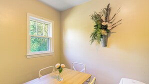 Un comedor con una mesa de madera, sillas blancas, un florero con flores, un ventanal y una jardinera colgante.