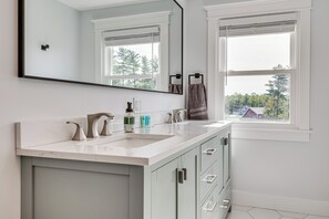 A modern bathroom with a white vanity, two sinks, a large mirror, and a window with a view of trees and buildings. - Golf, Hike & Explore: Family Home in Pembroke! (Suncook)
