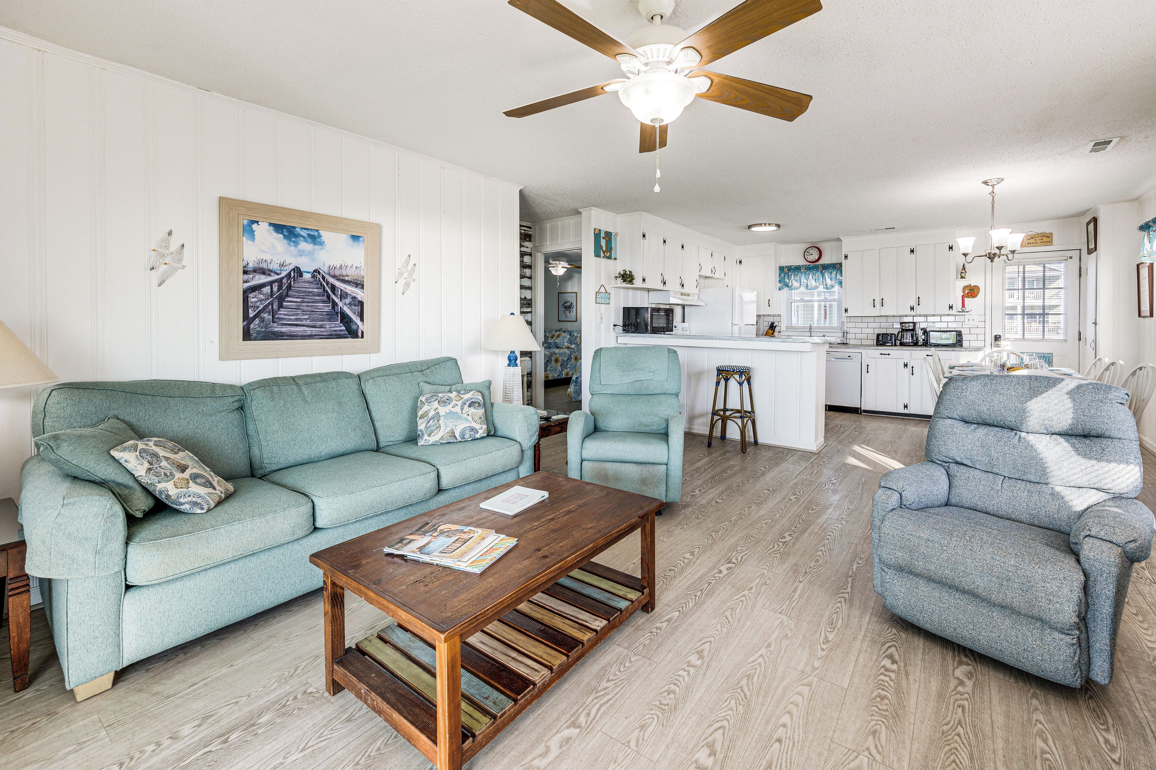 A living room with a sofa, armchair, and coffee table, a kitchen in the background, and a ceiling fan.