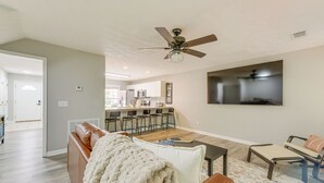 A modern living room with a brown leather sofa, a white throw blanket, and a coffee table. There is a ceiling fan and a flat-screen TV mounted on the wall. - 2 Mi to Tiger Territory! Condo w/ Community Pool (Auburn)