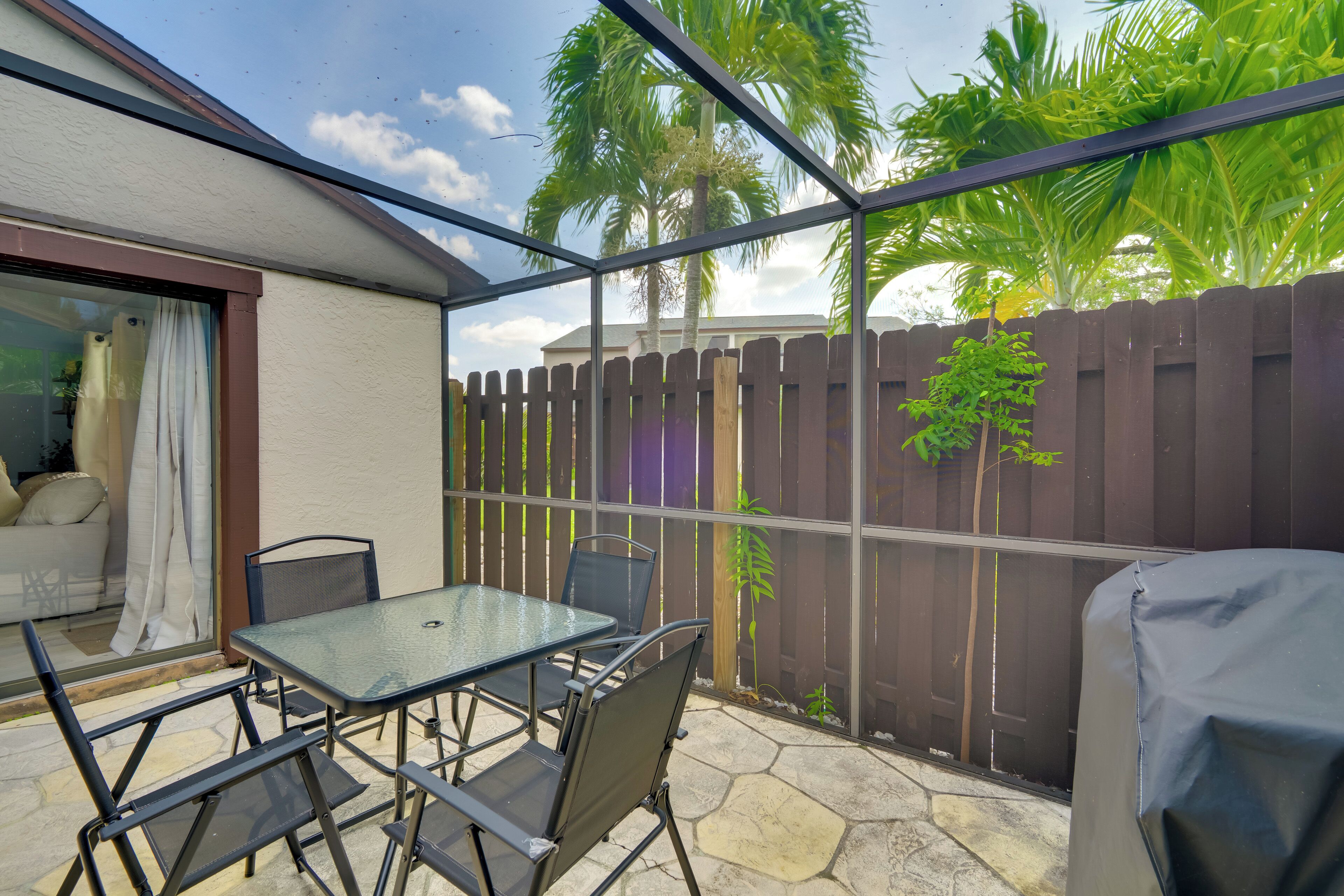 A covered patio with a glass table and chairs, a wooden fence, and a view of palm trees.