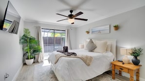 A bedroom with a bed, a ceiling fan, a nightstand, a potted plant, and a view of the outdoors.